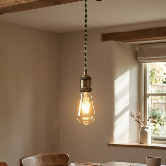 A rustic dining room scene with a wooden table set for dinner under a hanging Edison-style bulb pendant light with a green cord. The room has exposed wooden beams, cream walls, and a window with natural light.