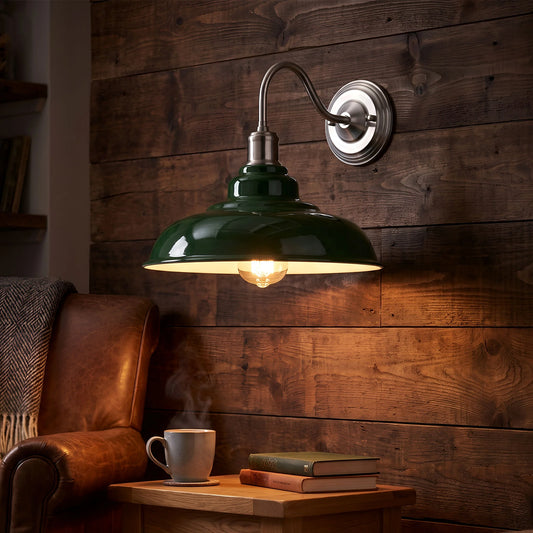 A green dome-shaped wall sconce with a silver metal wall mount and curved arm, illuminated with a warm light, shown mounted on a rustic dark wood plank wall above a wooden table with a mug and books.