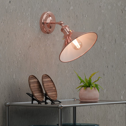 A rose gold industrial metal adjustable wall light sconce with a cone-shaped shade and a bulb, mounted on a grey wall.