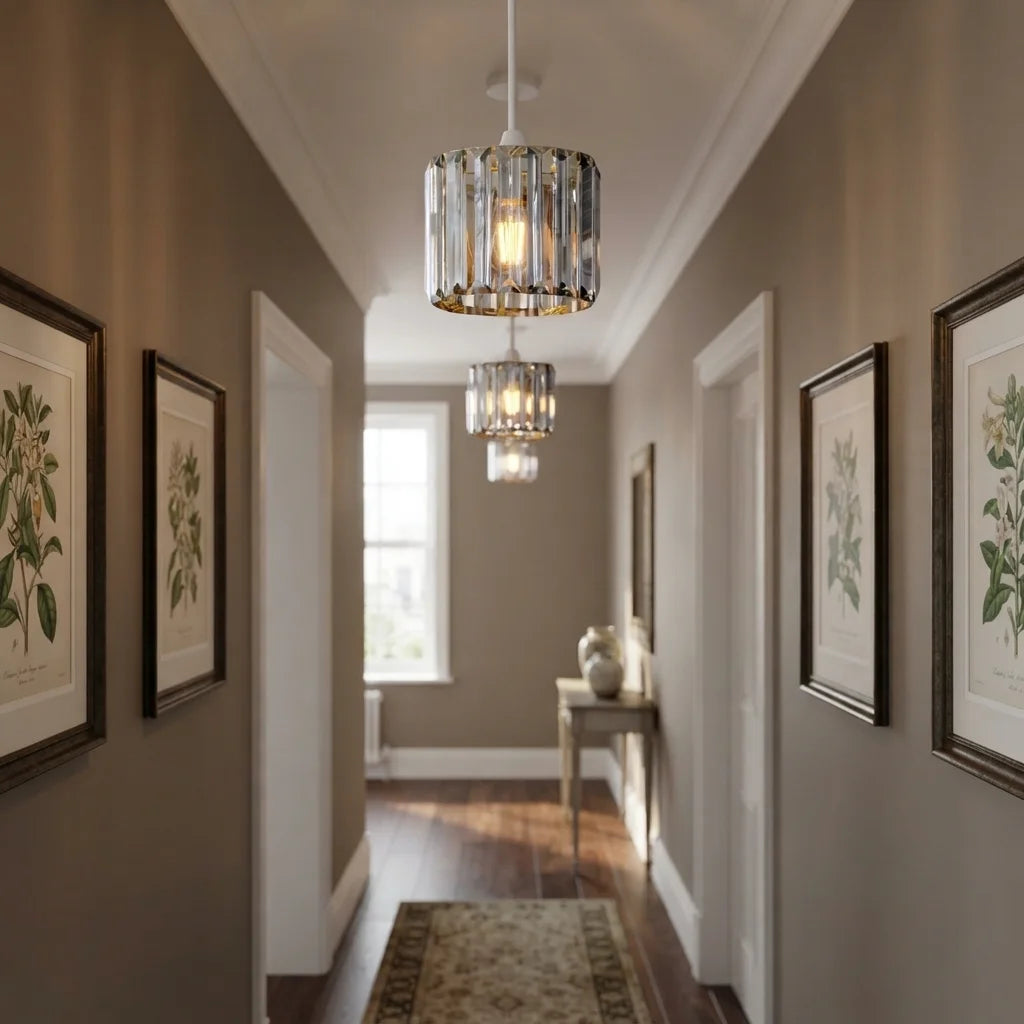 A modern cylindrical crystal chandelier with a warm glowing light hangs in a hallway with brown walls, framed botanical prints, and a wooden floor.