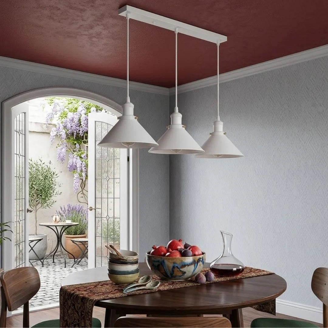 A modern three-light linear pendant chandelier with white conical shades hangs over a wooden dining table in a styled dining room with a dark red ceiling and light gray walls.