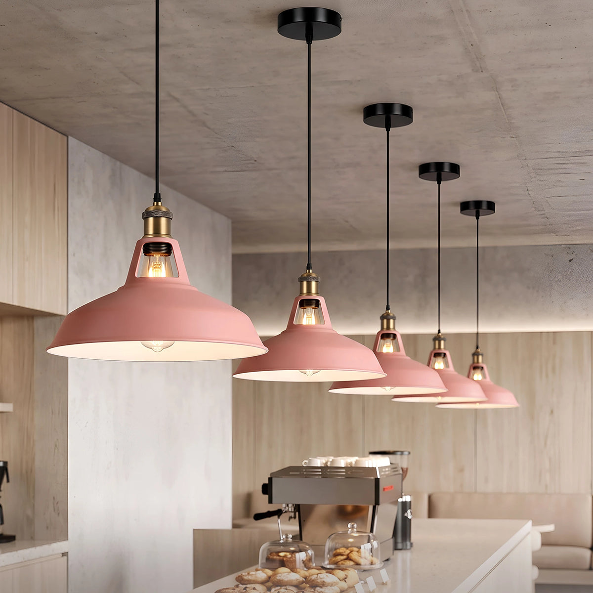 A row of modern pendant lights with dusty pink metal shades and brass fixtures hanging over a cafe counter in a concrete-walled interior.