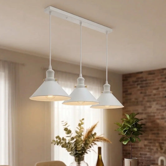 A three-light linear ceiling fixture with white downrod and three white conical pendant shades, shown hanging in a softly lit dining or living room with curtains, a brick wall, and plants in the background.