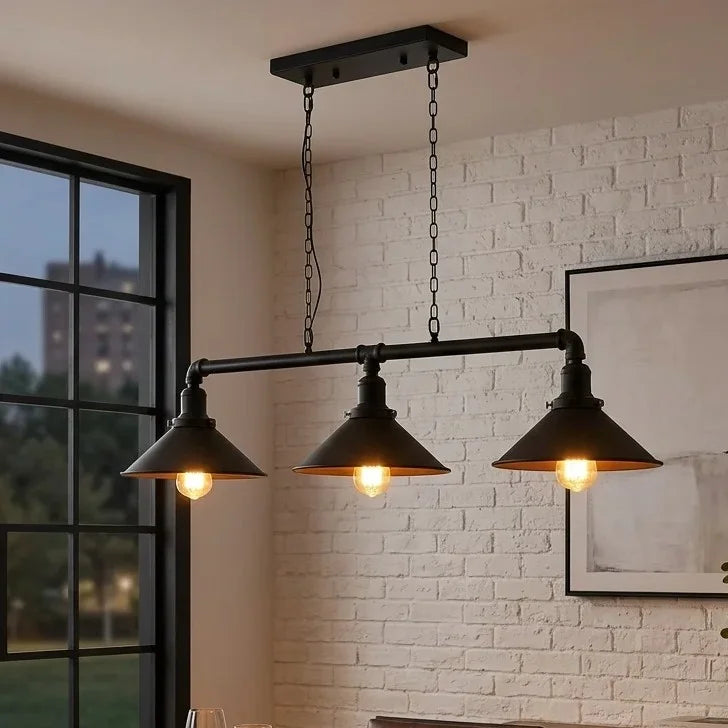 A three-light industrial pendant chandelier with a matte black horizontal bar, three conical shades, and visible warm glowing bulbs, hanging in a room with a white brick wall and window.