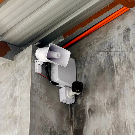 White PA horn speaker and CCTV security camera mounted on a concrete wall alongside a weatherproof junction box.