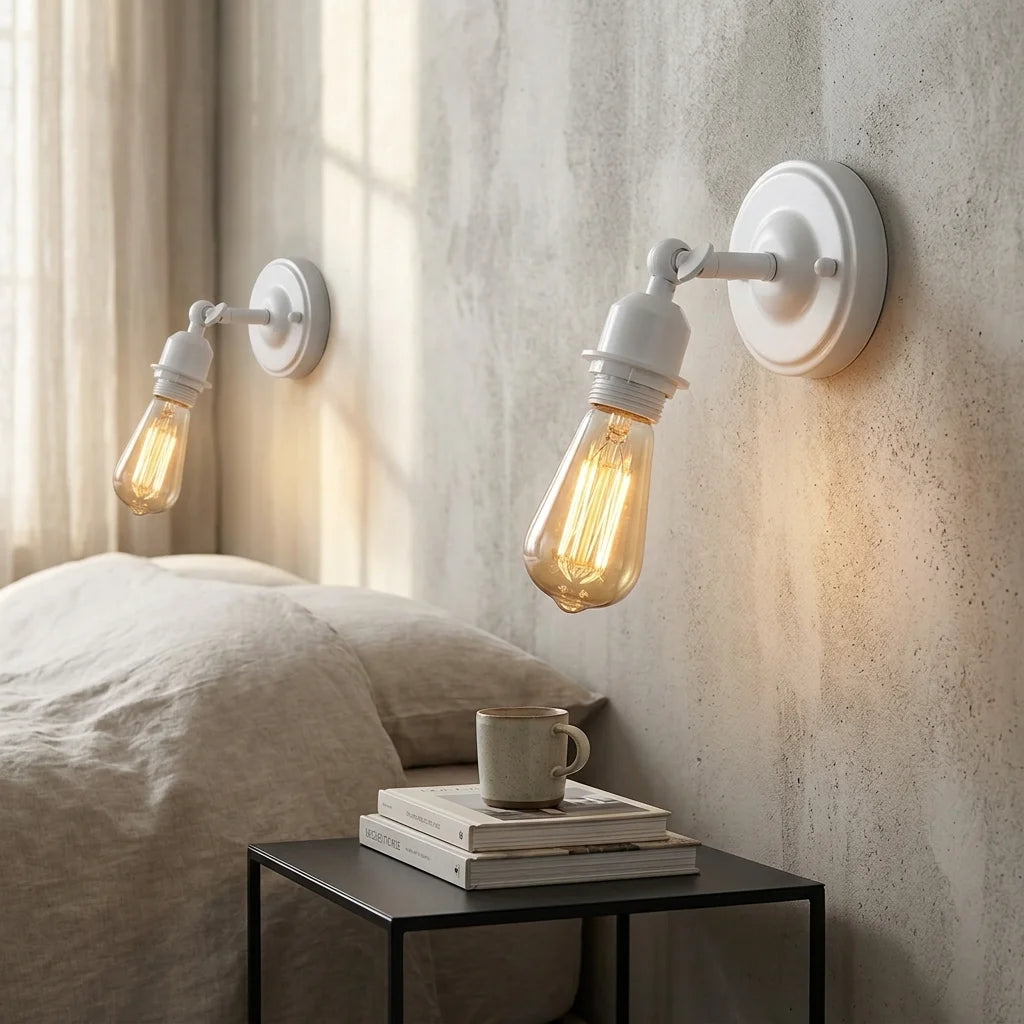 Two white industrial-style wall sconces with clear glass Edison bulbs mounted on a textured beige wall above a bed, with a small black side table and stacked books in the foreground.