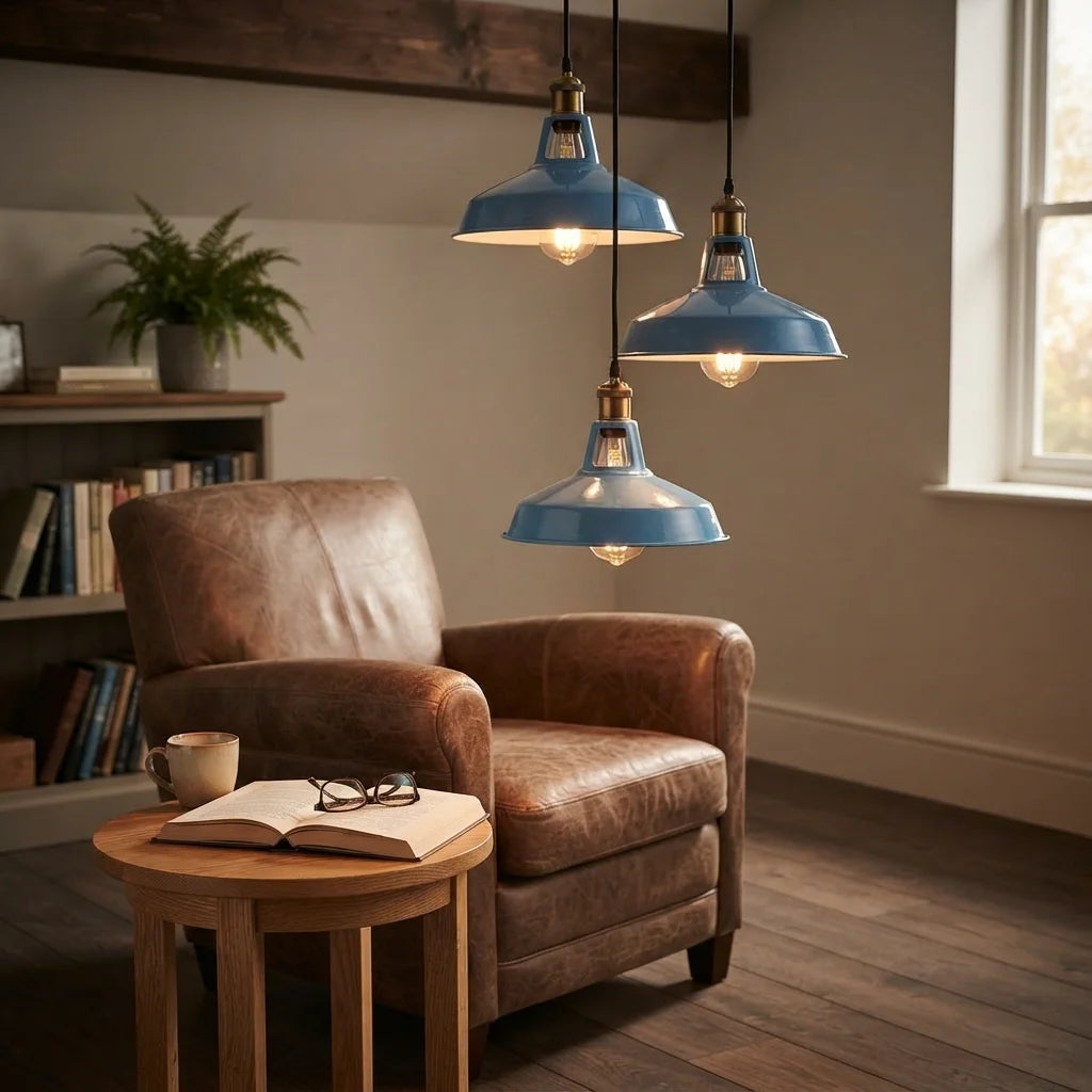 Three blue metal pendant lights with brass hardware hang over a brown leather armchair in a cozy, rustic living room setting with a wooden side table and bookshelf.