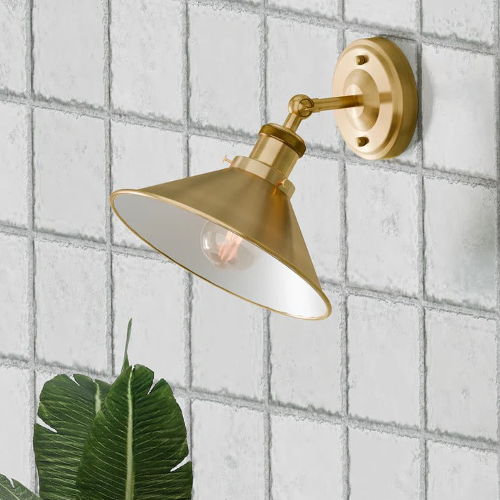  yellow brass-coloured industrial metal wall light sconce with an adjustable arm and a cone-shaped shade, mounted on a white tiled wall